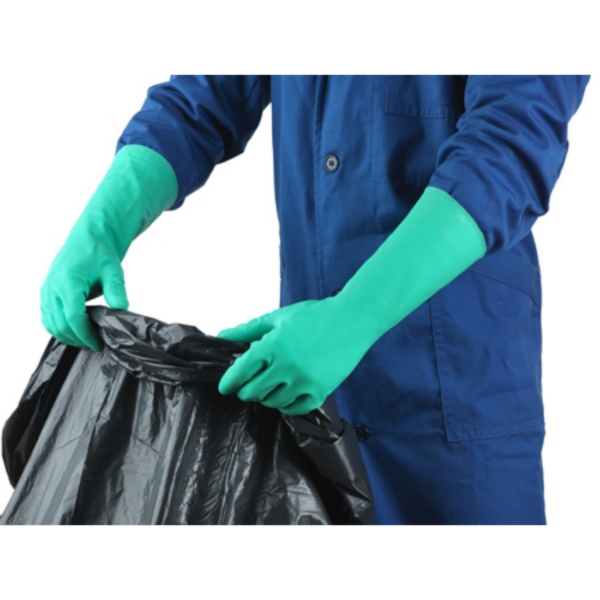 A man in blue overalls and green gloves holds a black garbage bag, ready for disposal.